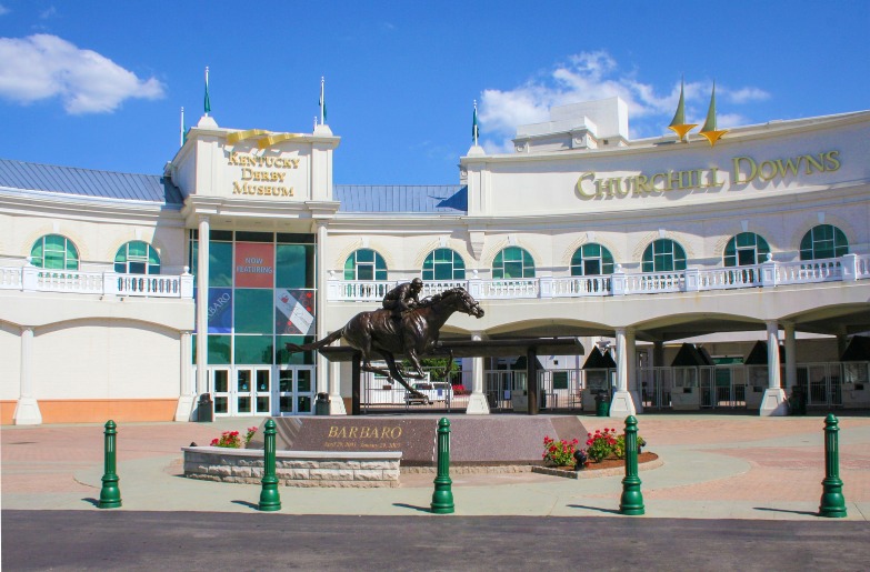 Standbeeld van een jockey te paard voor de sierlijke gevel van Churchill Downs en het Kentucky Derby Museum onder een blauwe lucht
