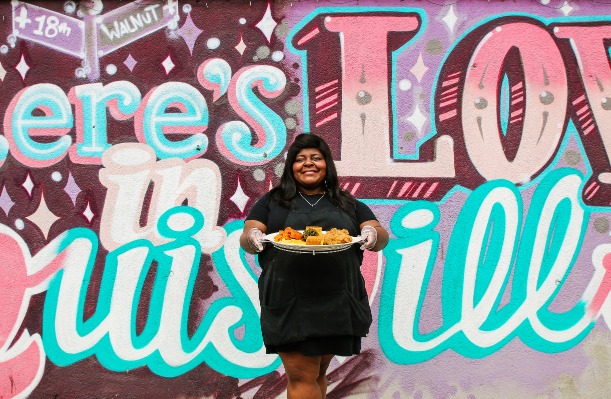 Vrouw houdt een bord met soul food vast voor een felgekleurde muurschildering met de tekst er is liefde in Louisville