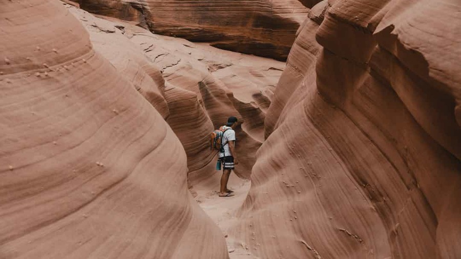 Persoon met rugzak loopt door een smalle kloof met gladde gegolfde roze en oranje rotswanden in Lower Antelope Canyon