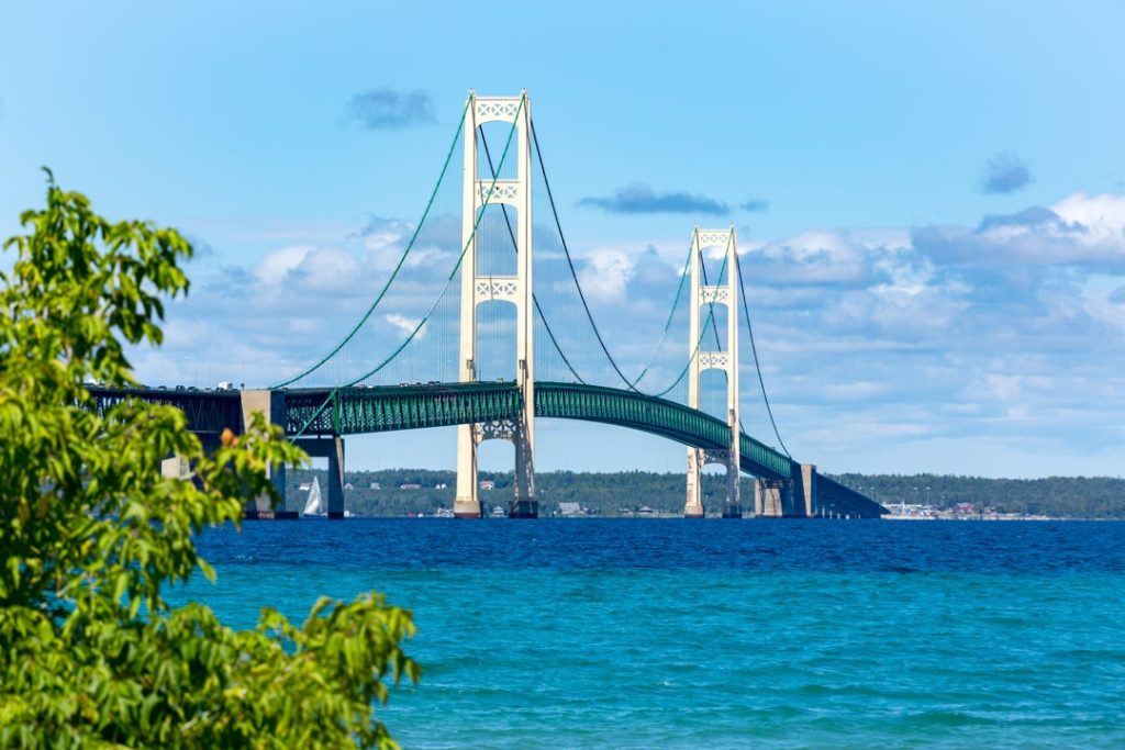 Hangbrug Mackinac Bridge met witte pylonen boven turquoise water, gezien tussen groene bladeren door