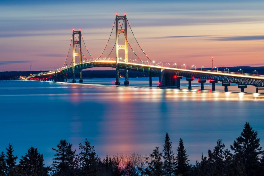 Verlichte hangbrug Mackinac Bridge met hoge pylonen weerspiegeld in het kalme water bij zonsondergang, dennenbomen op de voorgrond
