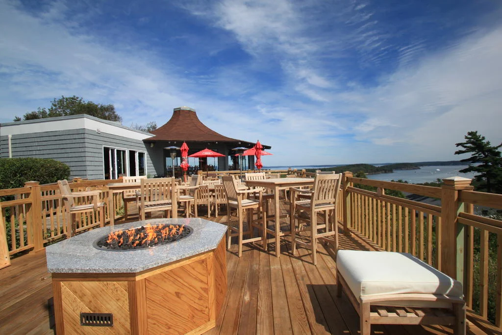 Houten terras met een vuurplaats, tafels en parasols met uitzicht over een baai onder een lichtbewolkte hemel in Maine