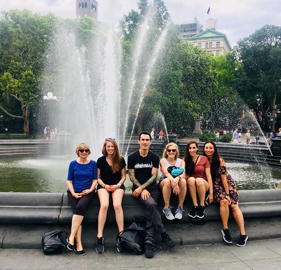 Groep reizigers zit op de rand van de fontein in Washington Square Park met opspattend water en een boogpoort op de achtergrond in Manhattan