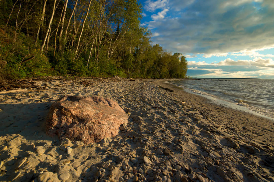 Stenig strand met een grote roze rots op de voorgrond, berkenbomen langs de oever en een meer met golven in Manitoba