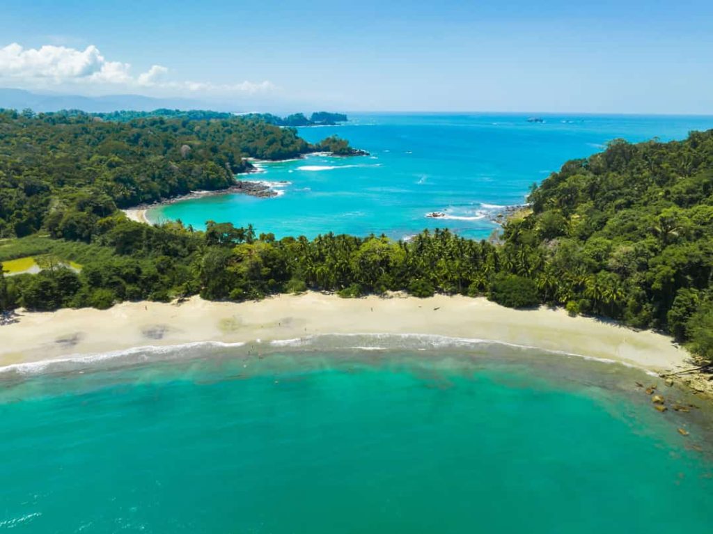Luchtfoto van een gebogen baai met wit zandstrand, palmbomen en helder turquoise water bij Manuel Antonio