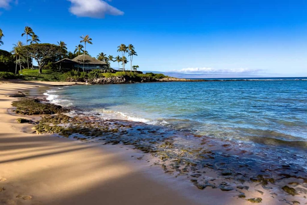 Rustig zandstrand met palmbomen en een huis op de rotsen, met kalm blauw water van de baai bij Kapalua op Maui