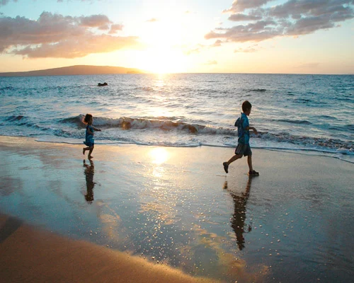 Twee kinderen rennen langs de waterlijn op een strand terwijl de zon net boven de horizon staat op Maui