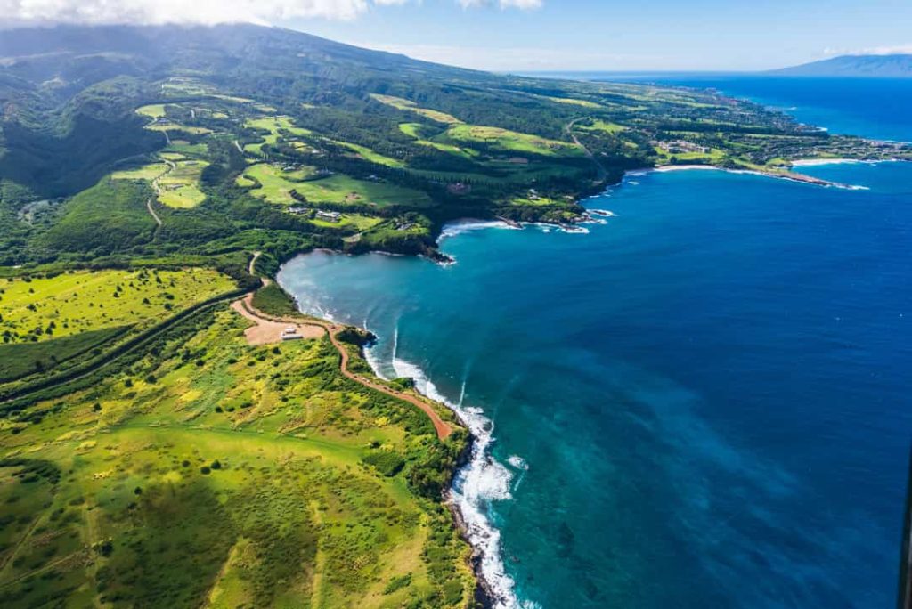 Luchtfoto van glooiende groene velden en een kronkelende kustweg langs een baai met helderblauw water op Maui