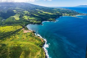 Luchtfoto van glooiende groene velden en een kronkelende kustweg langs een baai met helderblauw water op Maui