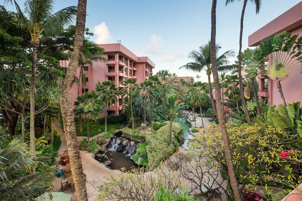 Uitzicht over een roze resort op Maui met een tropische tuin, waterval en hoge palmbomen