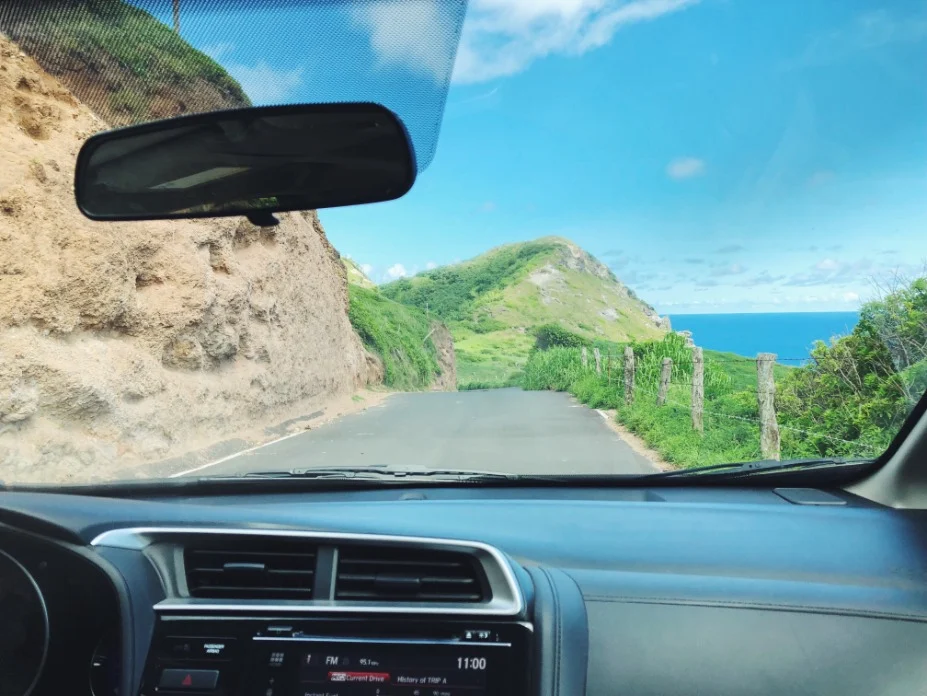 Blik door de voorruit van een auto op een smalle kustweg met groene heuvels en zee op Maui