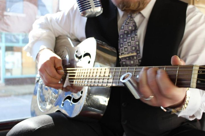 Close-up van de handen van een muzikant in pak die een zilveren resonatorgitaar bespeelt voor een microfoon