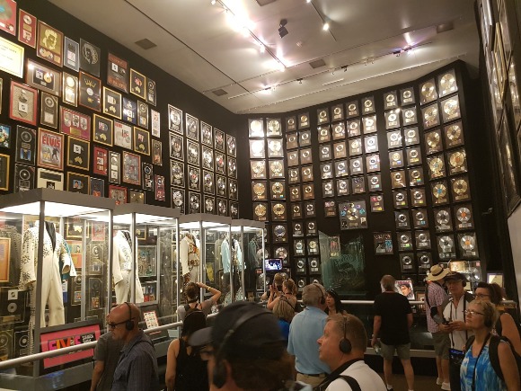Wand vol ingelijste gouden en platina platen en kostuums in vitrines in de trofeeënkamer van Graceland in Memphis, met bezoekers ervoor