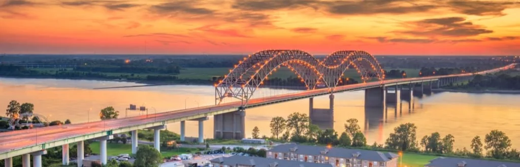 Verlichte boogbrug over de brede Mississippi rivier bij Memphis, gefotografeerd bij een gloeiende zonsondergang met appartementen aan de oever