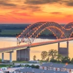 Verlichte boogbrug over de brede Mississippi rivier bij Memphis, gefotografeerd bij een gloeiende zonsondergang met appartementen aan de oever