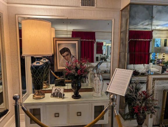Kamer met een geschilderd portret van Elvis Presley, een lamp en bloemen op een dressoir, weerspiegeld in spiegels met rode gordijnen