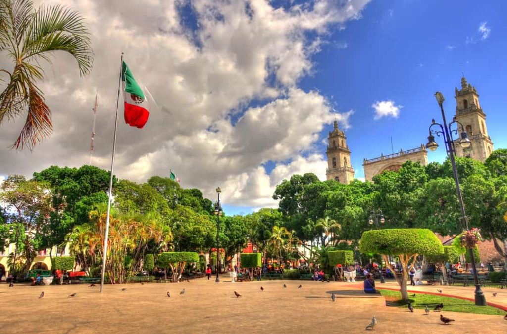 Een grote Mexicaanse vlag wappert op een plein met palmbomen, duiven en de torens van de kathedraal van Mérida op de achtergrond