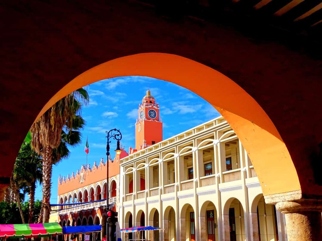 Door een grote oranje boog is het roodwitte stadhuis van Mérida met klokkentoren en zuilengang te zien