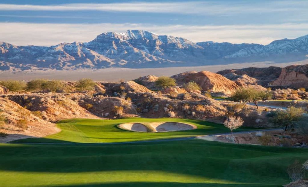 Groene golfgreen omringd door zandbunkers en rotsheuvels, met besneeuwde bergtoppen op de achtergrond bij Mesquite, Nevada
