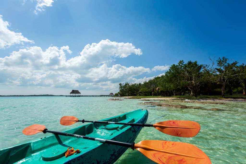 Vanuit een kajak met oranje peddels kijk je uit over glashelder turquoise water met een steiger in de verte