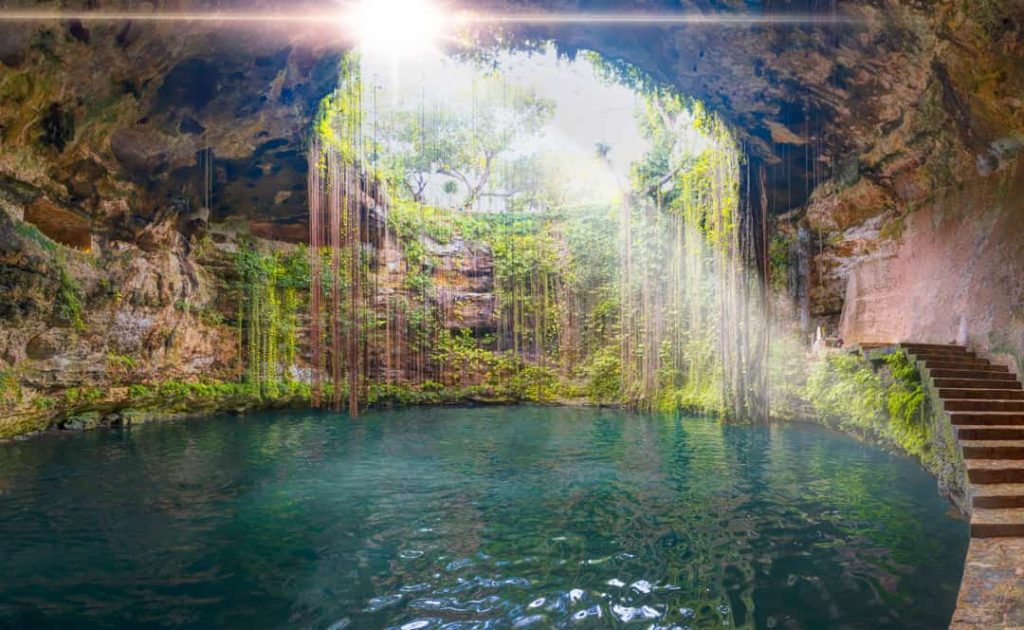 Zonlicht valt door een opening in een cenote in Yucatán, Mexico, waar hangende wortels boven turquoise water afdalen