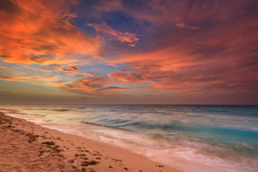 Oranje en rode wolkenlucht bij zonsondergang boven een zandstrand met turquoise golven in Mexico