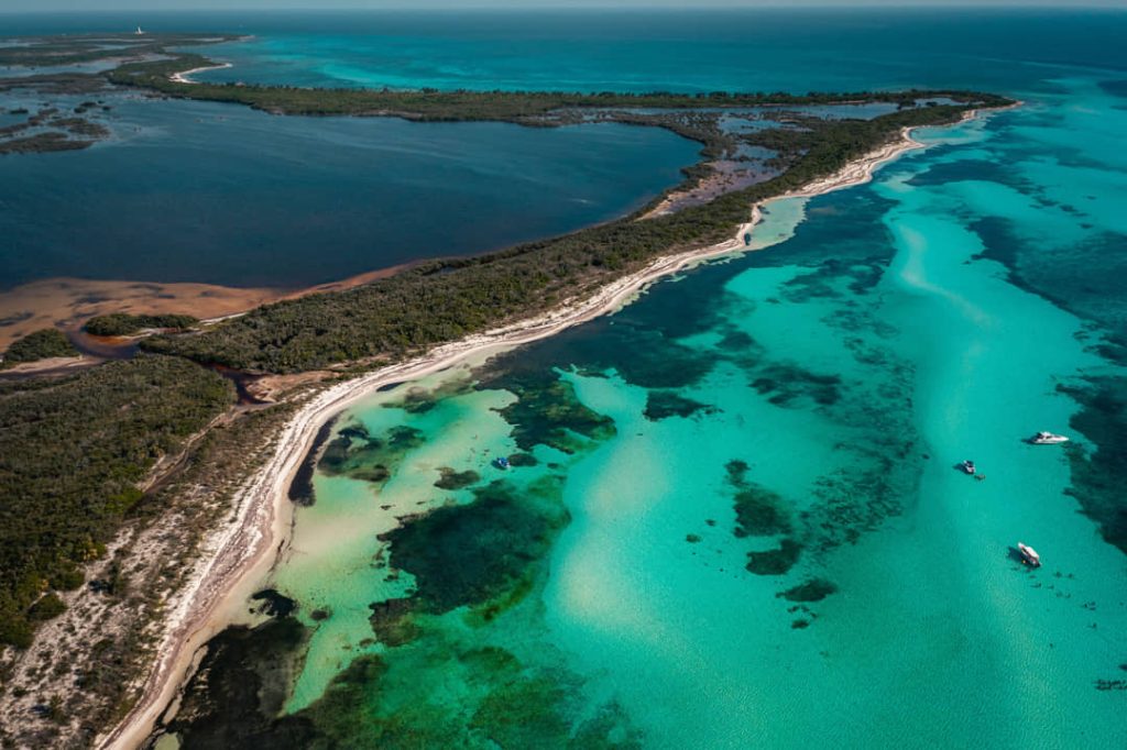 Luchtfoto van een smalle kustlijn met mangrovebos aan de ene kant en een koraalrif met bootjes aan de andere kant