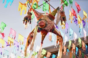 Een versierde piñata hangt tussen talloze kleurrijke papieren vlaggetjes tegen een lichte lucht