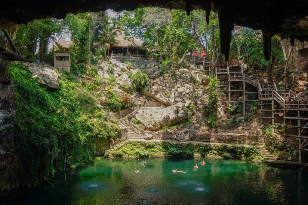 Mensen zwemmen in het heldere groene water van een cenote omringd door rotsen, begroeiing en houten trappen