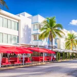 Zonnige straat met witte art-decogebouwen, palmbomen en rode parasols op terrasjes langs Ocean Drive in Miami