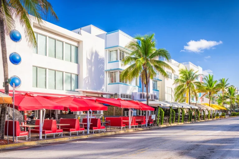 Zonnige straat met witte art-decogebouwen, palmbomen en rode parasols op terrasjes langs Ocean Drive in Miami