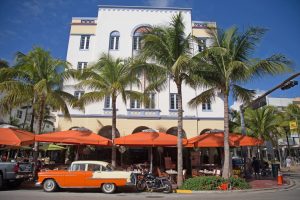 Wit art-decogebouw met oranje parasols op het terras en een oranje klassieke auto geparkeerd tussen palmbomen aan Ocean Drive