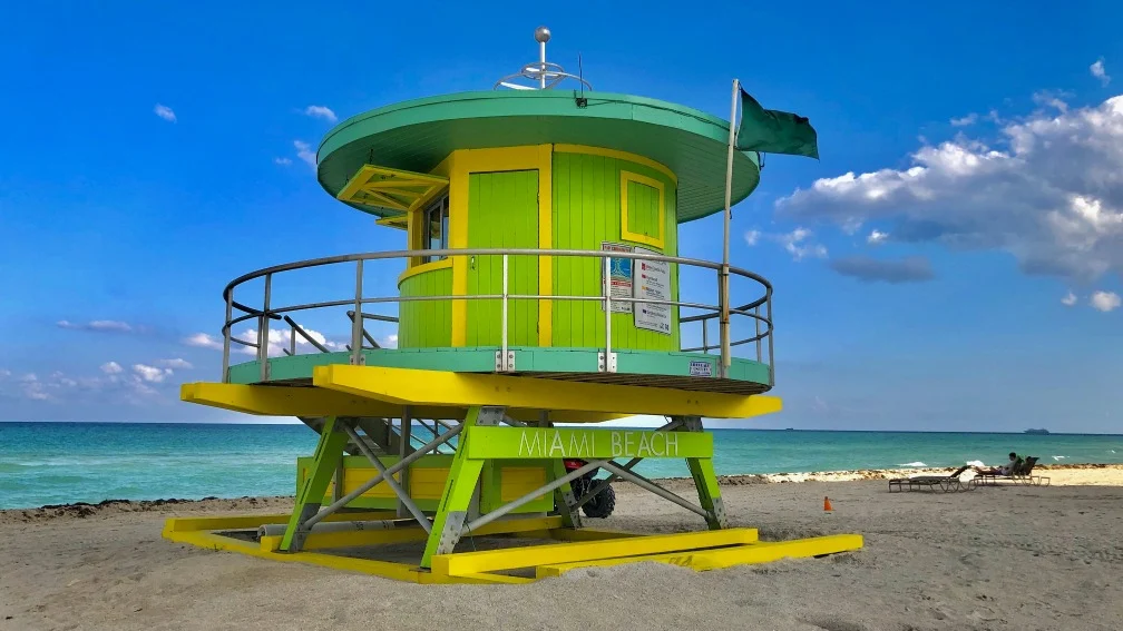 Ronde reddingstoren in groen en geel met de tekst Miami Beach op een leeg zandstrand onder een blauwe lucht