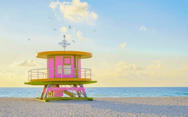 Een roze met geel reddingspostje met opschrift Miami Beach staat op een leeg zandstrand terwijl vogels overvliegen