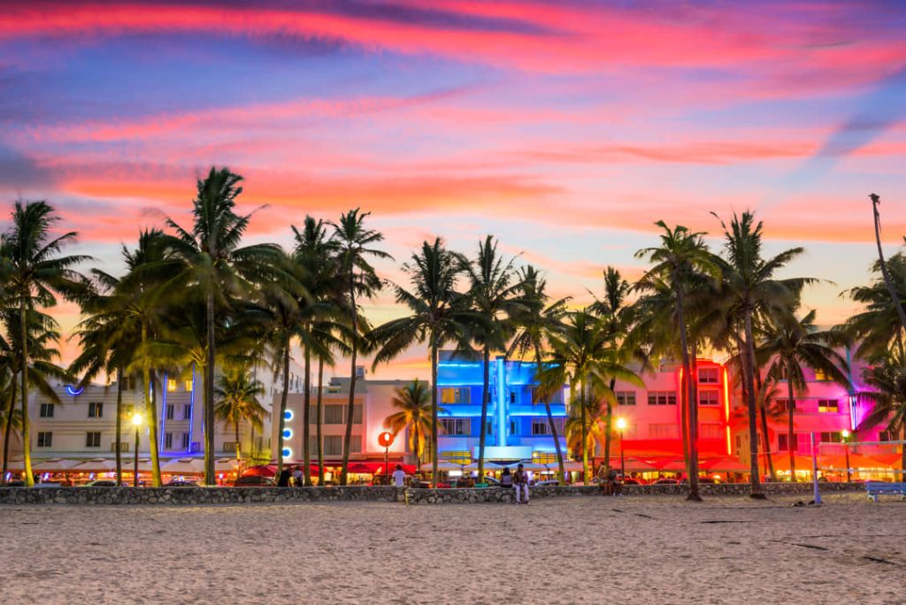 Palmbomen langs Ocean Drive in Miami Beach met neonverlichte art deco hotels onder een roze en paarse avondlucht