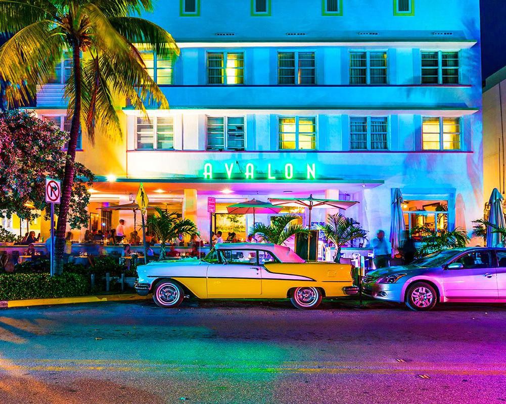 Verlicht art deco hotel Avalon aan Ocean Drive in Miami Beach met een gele klassieke cabriolet ervoor geparkeerd in de avond