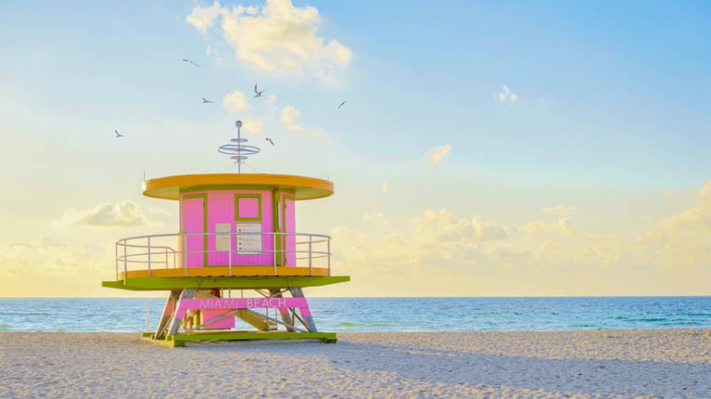 Kleurrijke gele en roze reddingstoren met de tekst Miami Beach op een leeg zandstrand onder een blauwe lucht met vogels