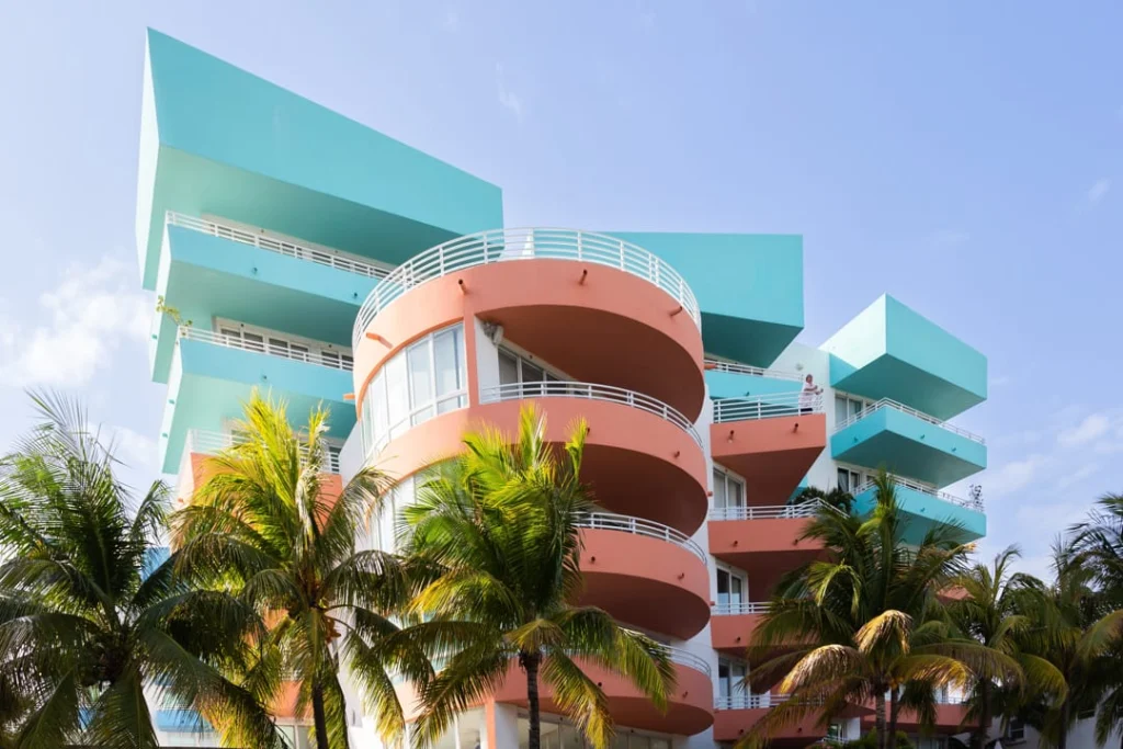 Modern art deco gebouw in turkoois en koraalroze met ronde balkons, omringd door palmbomen onder een blauwe hemel in Miami Beach