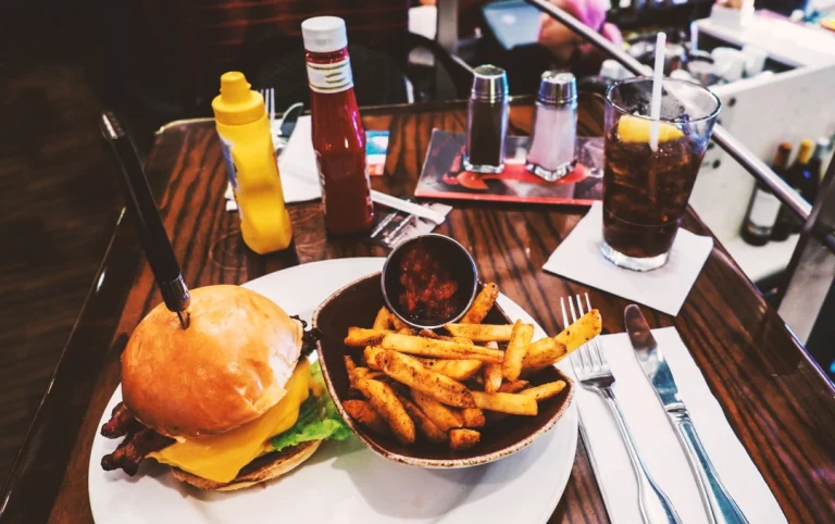 Een hamburger met bacon en kaas naast een bakje kruidige friet en cola op een houten tafel in een Amerikaans diner