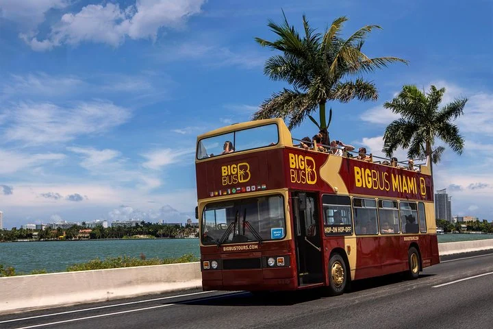 Een rode en gele Big Bus dubbeldekker rijdt over een brug met palmbomen en de skyline van Miami op de achtergrond