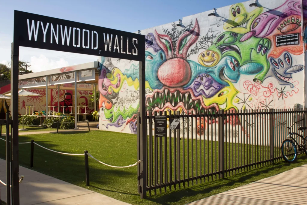 Zwart bord met opschrift Wynwood Walls bij de ingang van het street art park in Miami, met een kleurrijke muurschildering van fantasiefiguren op de achtergrond