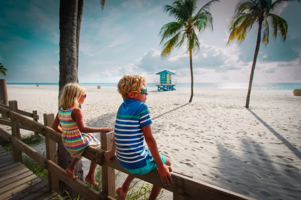 Twee kinderen zitten op een houten hek en kijken uit over het witte strand met palmbomen en een gele reddingspost