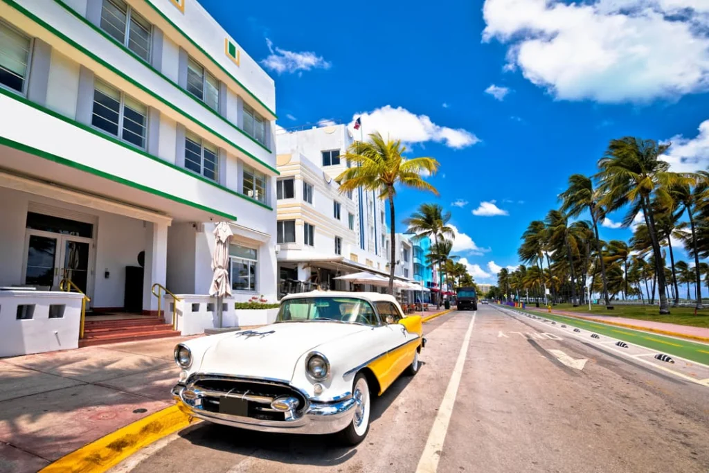 Een witgele klassieke auto staat geparkeerd voor art-decogebouwen aan een palmbomenlaan in Miami Beach