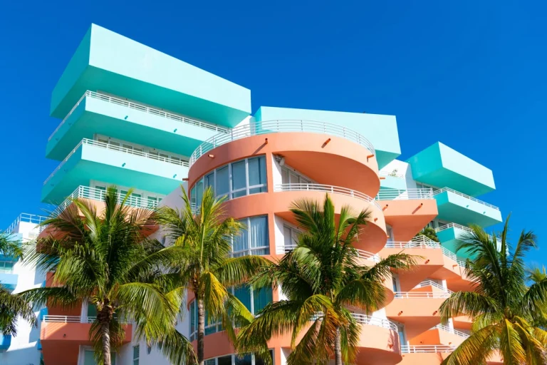 Kleurrijke art-decoflats in turquoise en oranje tinten steken af tegen een strakblauwe hemel, met palmbomen op de voorgrond in Miami