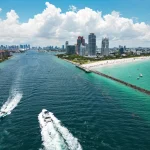 Luchtfoto van Miami met twee boten op het water, een pier en de skyline naast een turquoise strand
