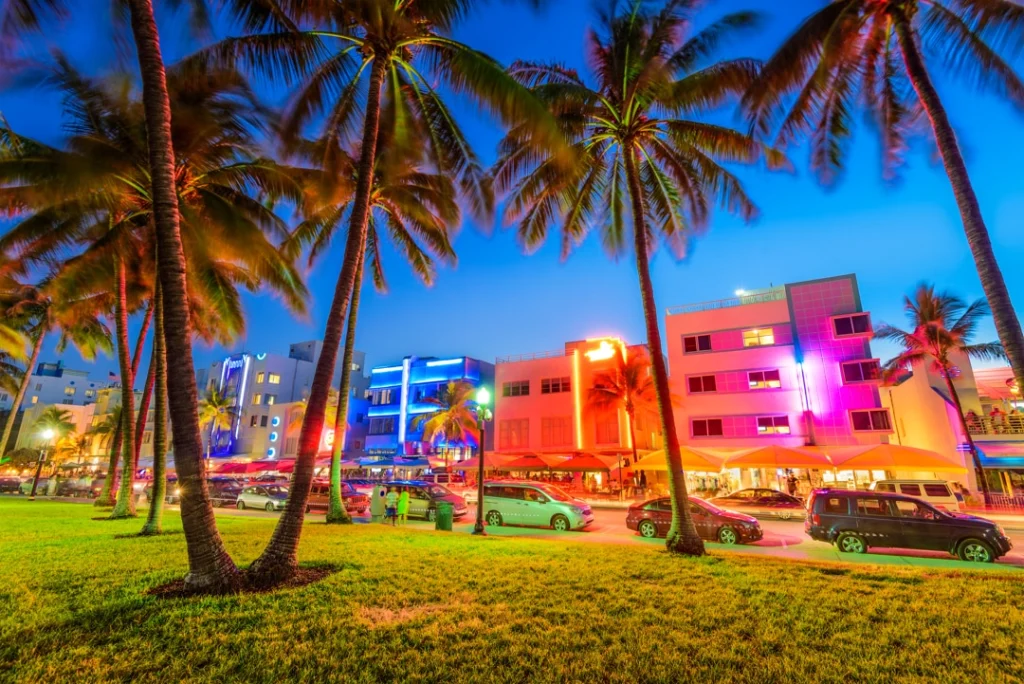 Palmbomen omlijsten de kleurrijk verlichte art-decogebouwen en drukke straat van Ocean Drive in Miami tijdens de avondschemering