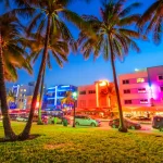 Palmbomen omlijsten de kleurrijk verlichte art-decogebouwen en drukke straat van Ocean Drive in Miami tijdens de avondschemering