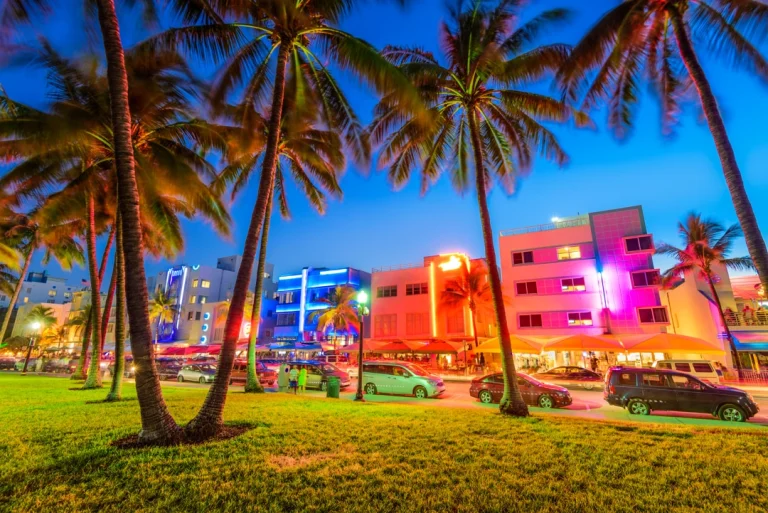 Palmbomen omlijsten de kleurrijk verlichte art-decogebouwen en drukke straat van Ocean Drive in Miami tijdens de avondschemering