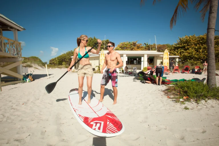 Een lachende vrouw met een peddel staat op een suppboard op het strand van Miami, terwijl een man ernaast toekijkt