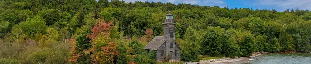 Verweerde houten vuurtoren met een klein huisje aan de oever, omgeven door dicht bos met herfstkleuren in Michigan